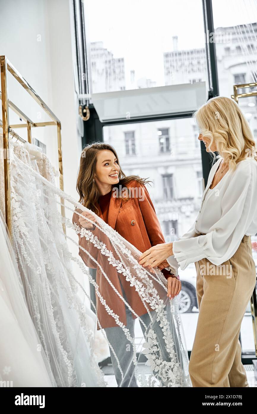 Une jeune belle mariée et sa mère examinent une délicate robe de mariée en dentelle sur un rack, envisageant la journée spéciale des mariées. Banque D'Images