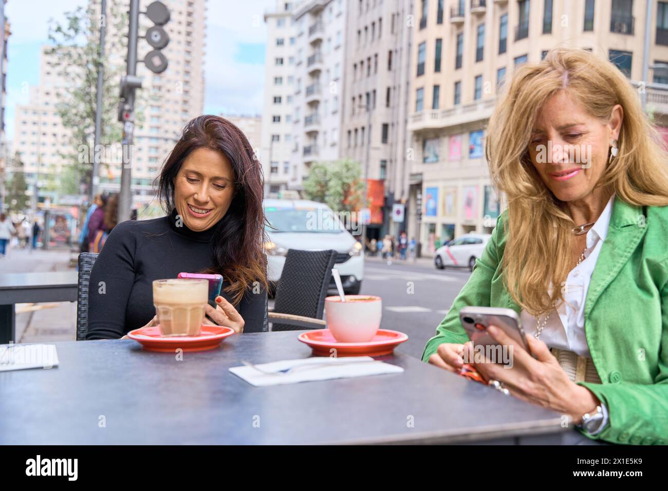 amies matures utilisant leurs smartphones assis sur une terrasse de bar prenant une tasse de café Banque D'Images