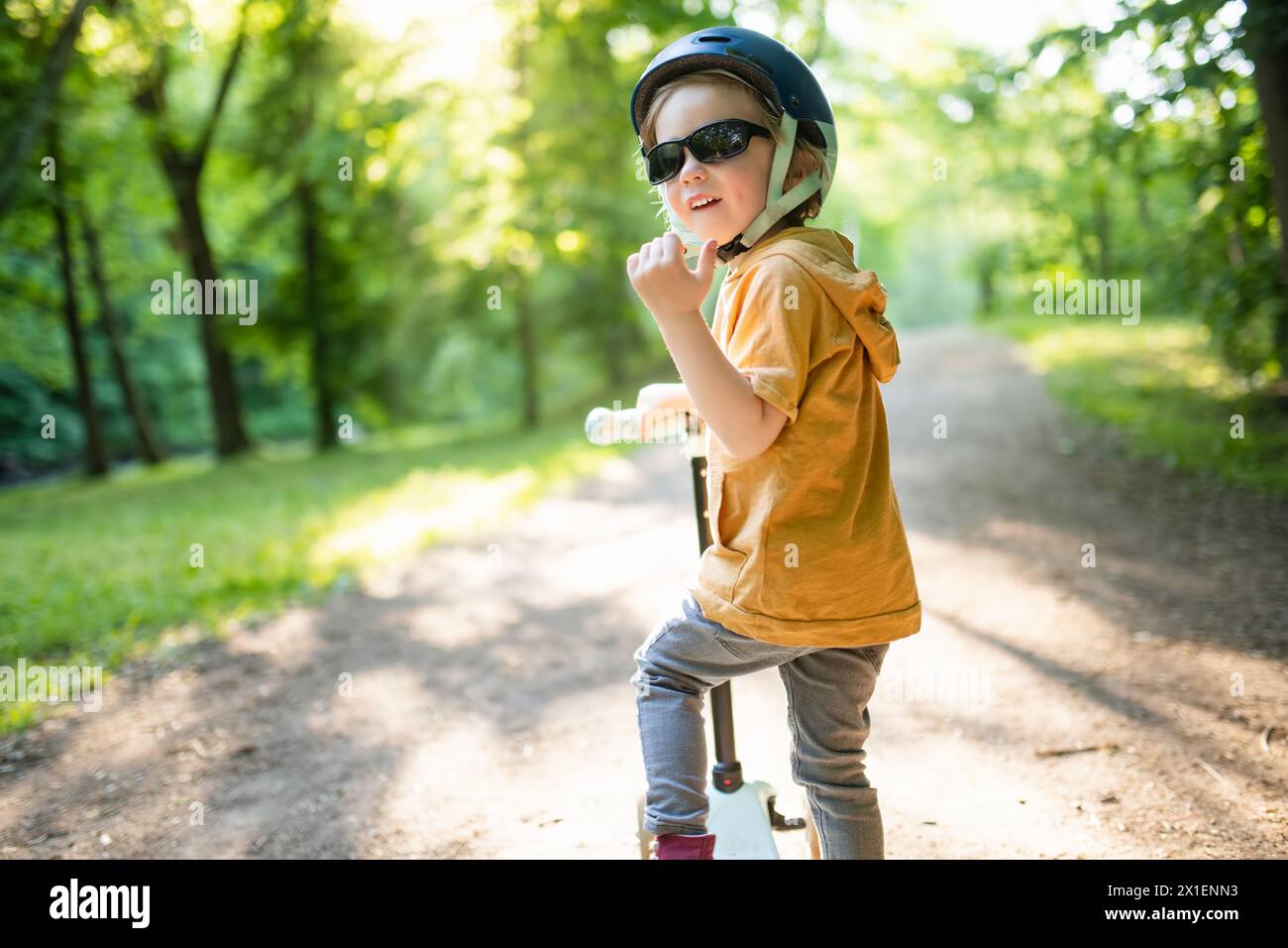 Adorable petit garçon chevauchant son scooter dans un parc de la ville le soir ensoleillé d'été. Jeune enfant chevauchant un rouleau avec un casque sur. Loisirs actifs et Outdoo Banque D'Images