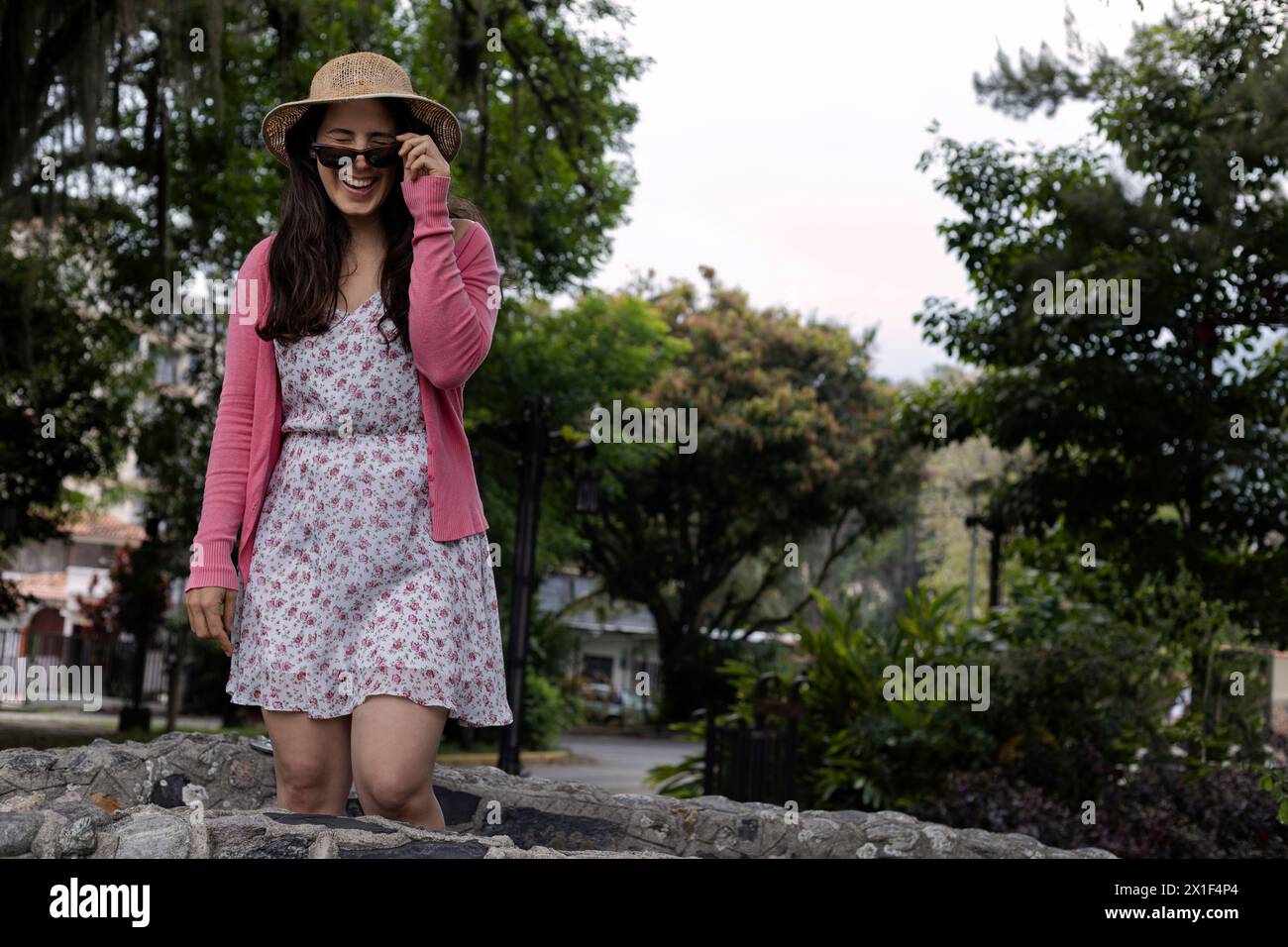 Jeune femme latino-américaine (33 ans) se tient avec une attitude positive regardant par-dessus ses lunettes. Porter un chapeau, une robe de fleur et un pull rose. Concept de ressort. Banque D'Images