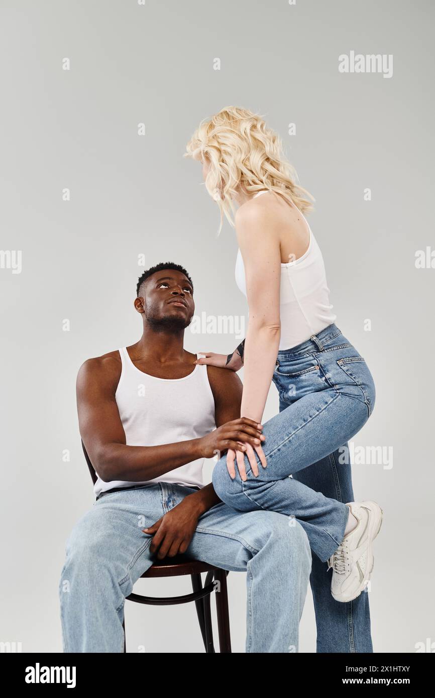 Un jeune couple interracial, l'homme assis sur une chaise à côté de la femme, exsudant l'unité et l'amour dans un studio sur un fond gris. Banque D'Images