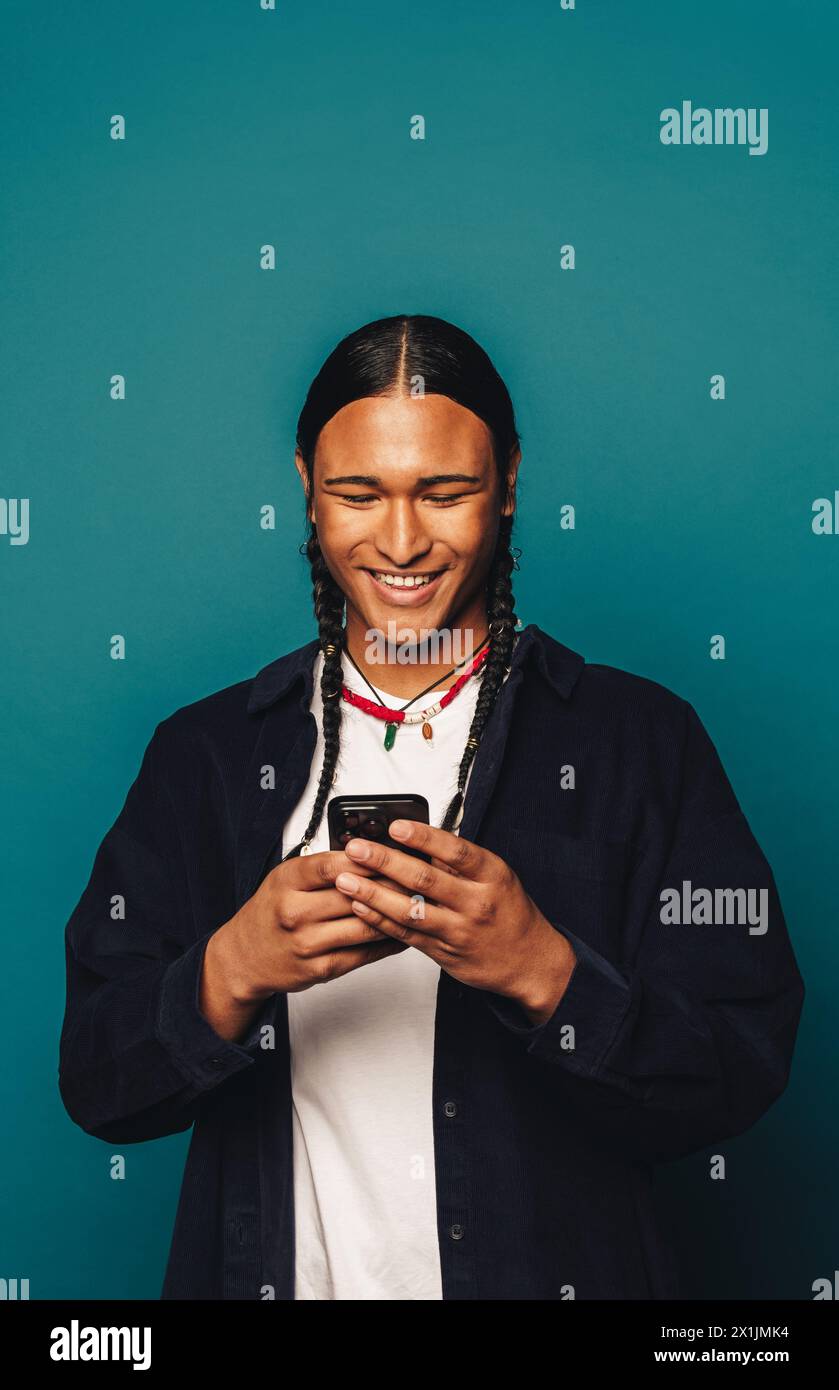 Homme heureux avec de longs cheveux tressés, portant des vêtements décontractés et un collier amérindien, debout dans un studio. Il sourit en lisant un message sur h. Banque D'Images