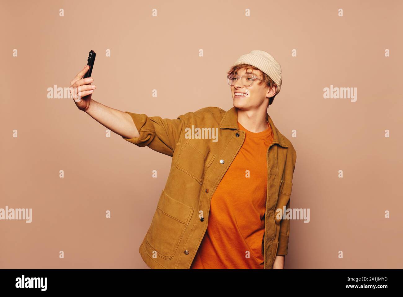 Homme souriant dans des vêtements décontractés se dresse contre un fond de pêche vibrant. Il tient un téléphone portable, prenant un selfie avec une expression heureuse. Banque D'Images