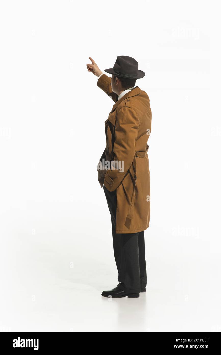 L'homme en trench-coat de style rétro et chapeau Fedora debout et pointant vers le haut isolent le fond blanc don. Banque D'Images