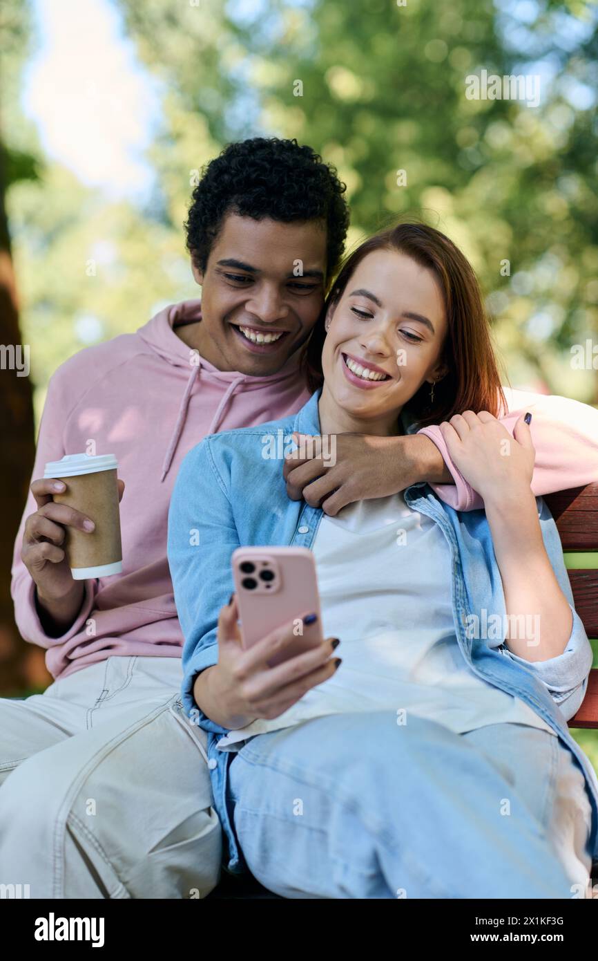 Couple diversifié dans une tenue vibrante assis sur le banc du parc, engrossed dans l'écran de téléphone portable. Banque D'Images