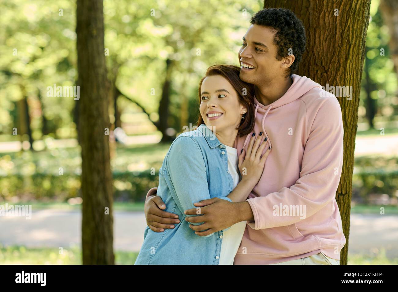 Un couple aimant et diversifié dans une tenue vibrante se tient ensemble à côté d'un arbre dans un parc. Banque D'Images