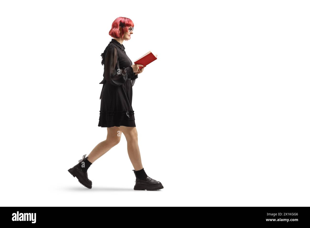Plan de profil complet d'une jeune femme aux cheveux roux dans une robe noire marchant et lisant un livre isolé sur fond blanc Banque D'Images