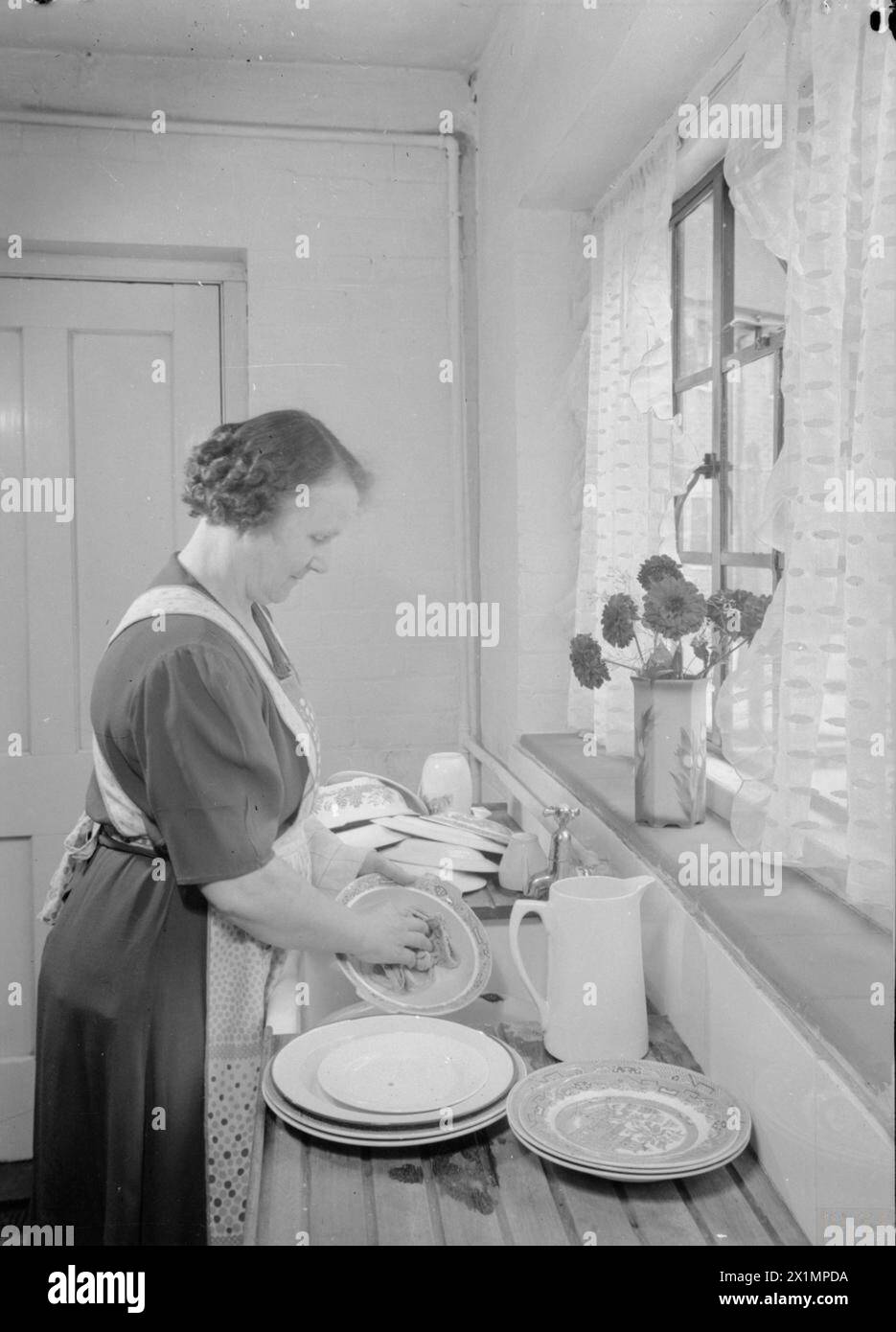 LES CHALETS DE FREEFOLK : LA VIE DANS LE VILLAGE DE FREEFOLK, HAMPSHIRE, ANGLETERRE, Royaume-Uni, 1943 - Mme Shadwell se tient devant le grand évier dans la cuisine de sa maison dans le village de Freefolk, Hampshire et lave la vaisselle. L'évier est placé sous la fenêtre, avec une grande planche de drainage des deux côtés de l'évier. Un vase de fleurs décore le rebord de la fenêtre, Banque D'Images