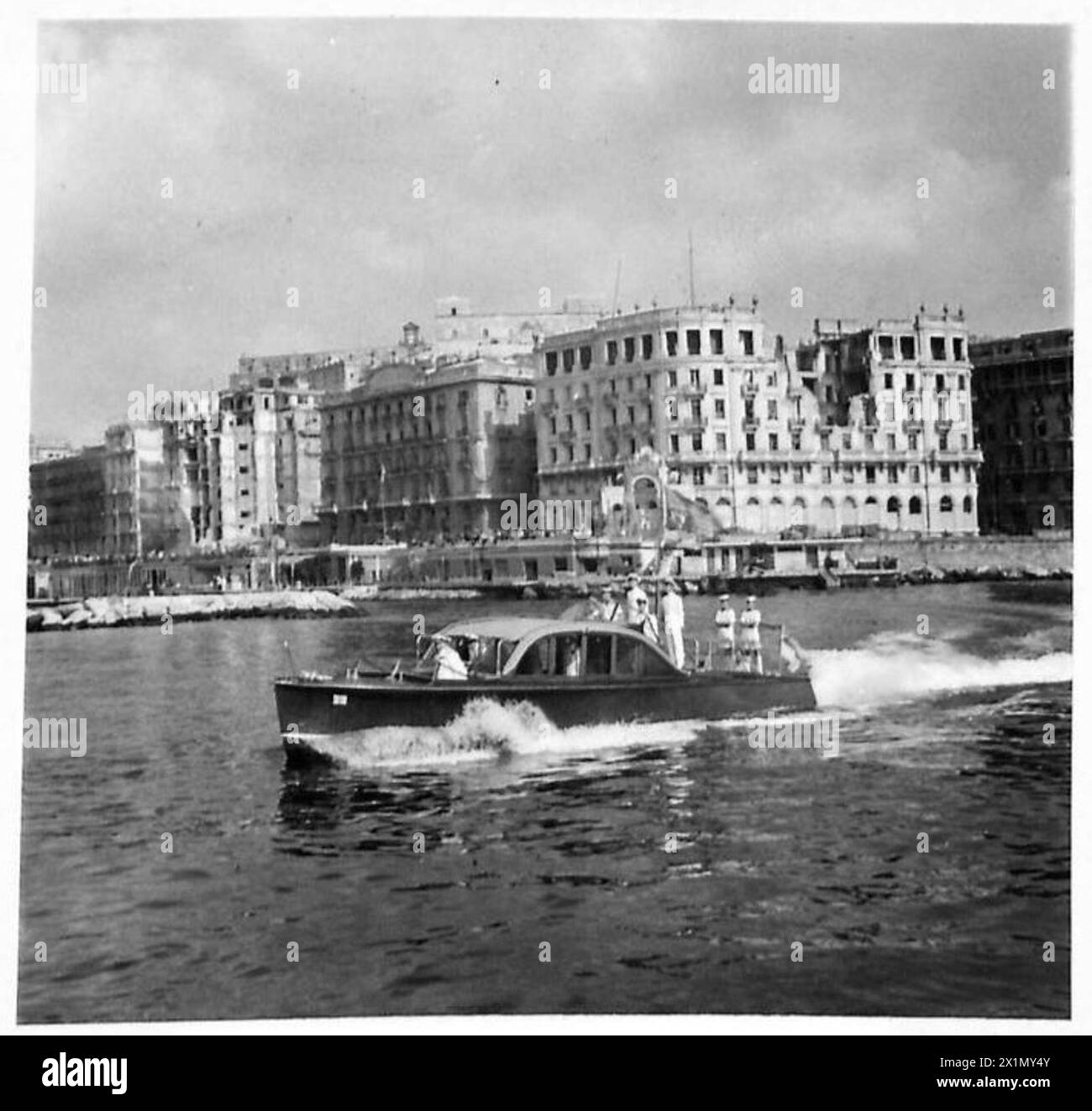 Sa Majesté le Roi part dans la péniche royale pour une visite des unités navales ancrées dans la baie de Naples, en Italie, avec des bâtiments de bord de mer marqués par des bombes en vue, l'armée britannique. Banque D'Images