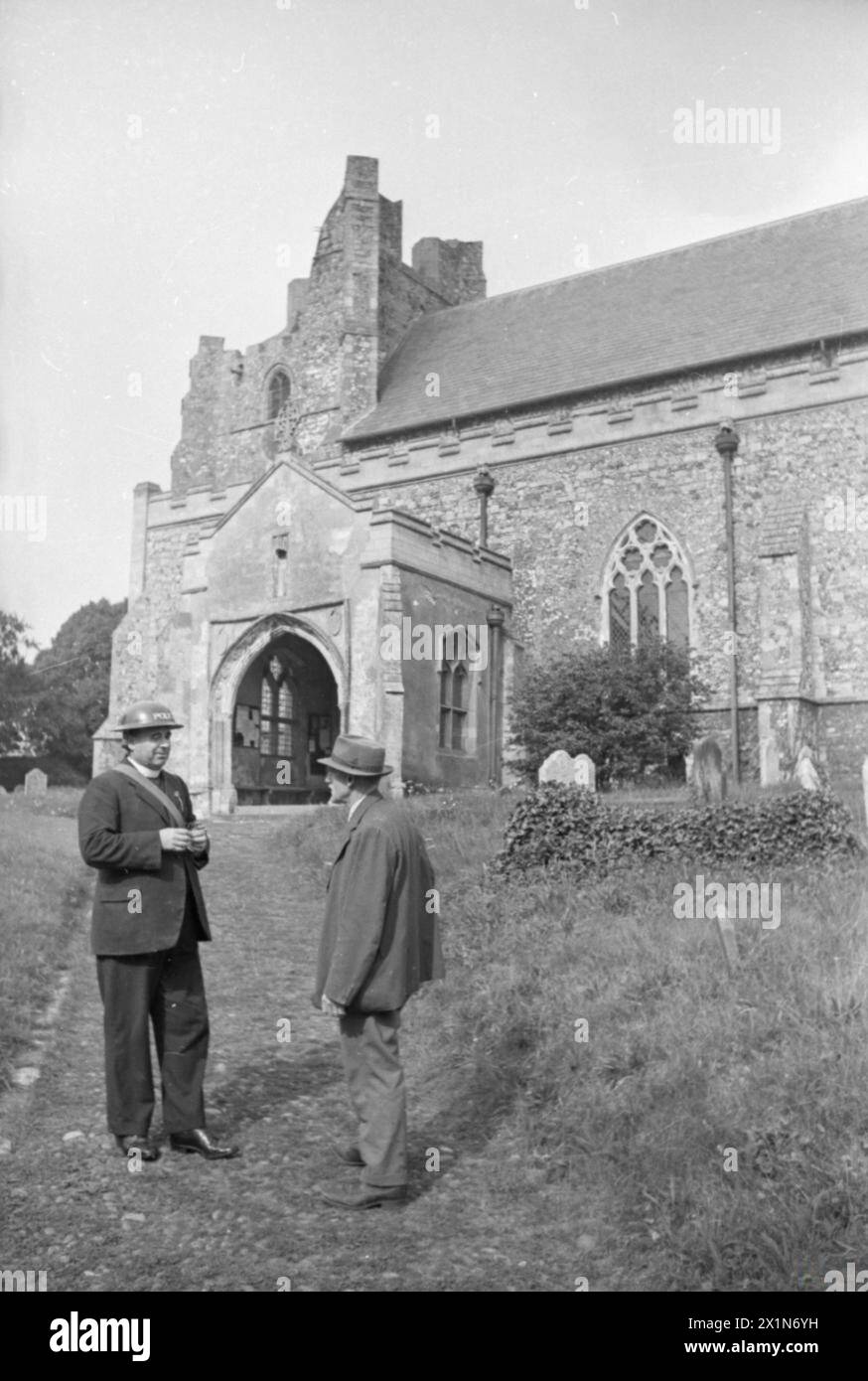 VILLAGE D'INVASION : LA VIE QUOTIDIENNE À ORFORD, SUFFOLK, ANGLETERRE, 1941 - le révérend A. C. Hopkins, le recteur de l'église Saint-Barthélemy, parle à un paroissien sur le chemin menant à l'église. Le révérend Hopkins a servi pendant la première Guerre mondiale et est maintenant le chef de la police spéciale du village. Avec son collier de chien, il porte un casque de police en acier et la sangle de son étui de masque à gaz peut également être vue par-dessus son épaule. Notez qu'une partie de la tour est manquante, après son effondrement en 1830, Banque D'Images