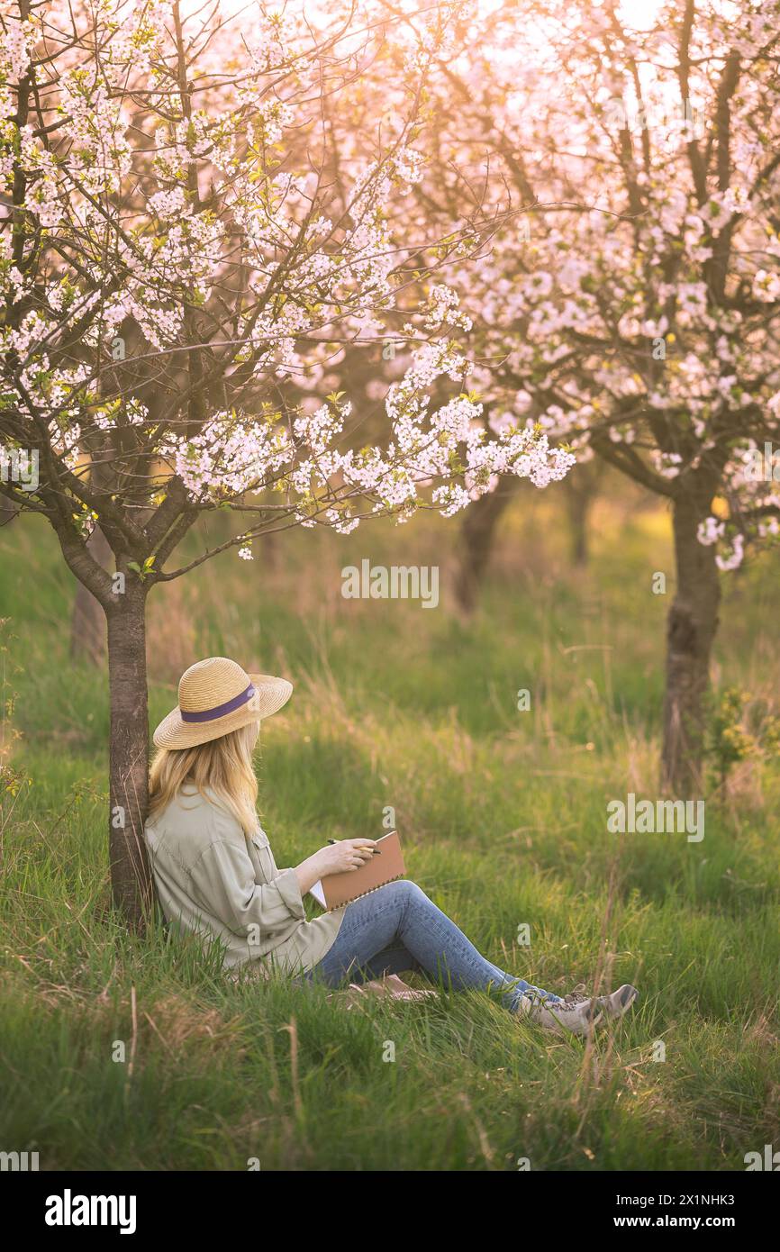 Écrivain cherche l'inspiration pour écrire son livre tout en contemplant dans le verger florissant de printemps. Femme écrivant dans un cahier Banque D'Images