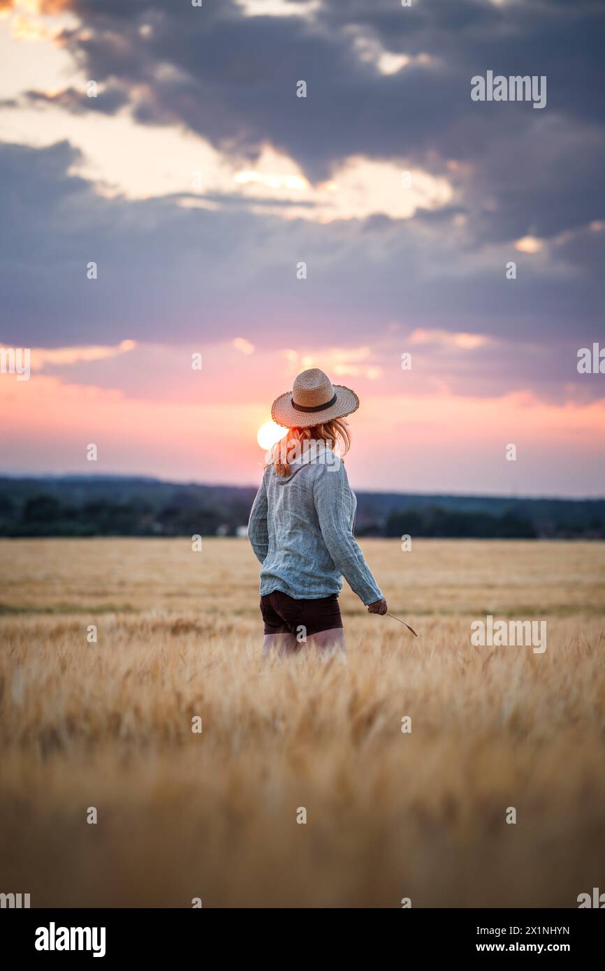Agricultrice satisfaite regarde le champ d'orge dans sa ferme. Femme debout dans un champ agricole pendant le coucher du soleil. Contrôle de la qualité du grain avant récolte Banque D'Images