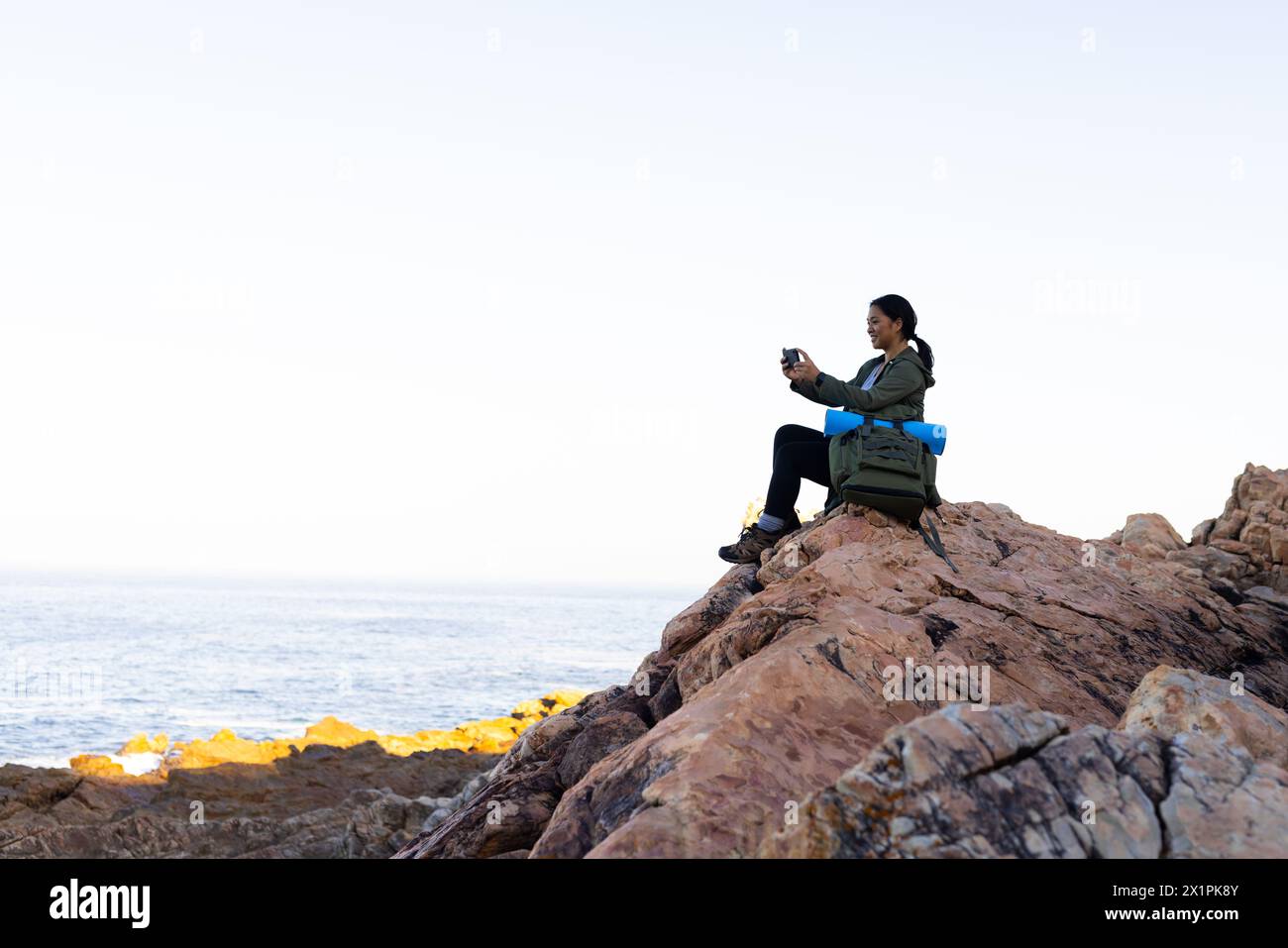 Randonneur féminin biracial assis sur des rochers au milieu de la nature, tenant un appareil photo et un smartphone, avec copie s. Banque D'Images