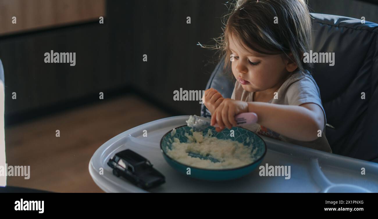 Petite fille dans la cuisine mangeant avec impatience du riz avec une cuillère indépendamment. Banque D'Images