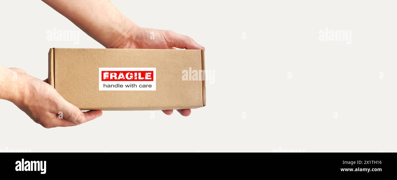 Une boîte en carton rectangulaire dans les mains pour l'emballage d'articles fragiles sur un fond clair Uni. Étiquette d'avertissement rouge, autocollant pour emballage « fragile » Banque D'Images