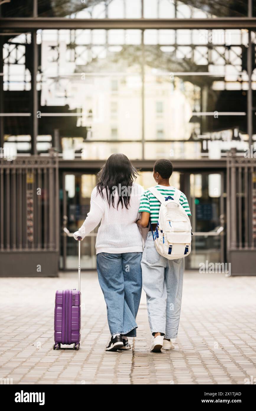 Vue arrière de deux jeunes femmes tirant une valise et marchant ensemble à travers une ville lors d'un voyage de week-end. Banque D'Images