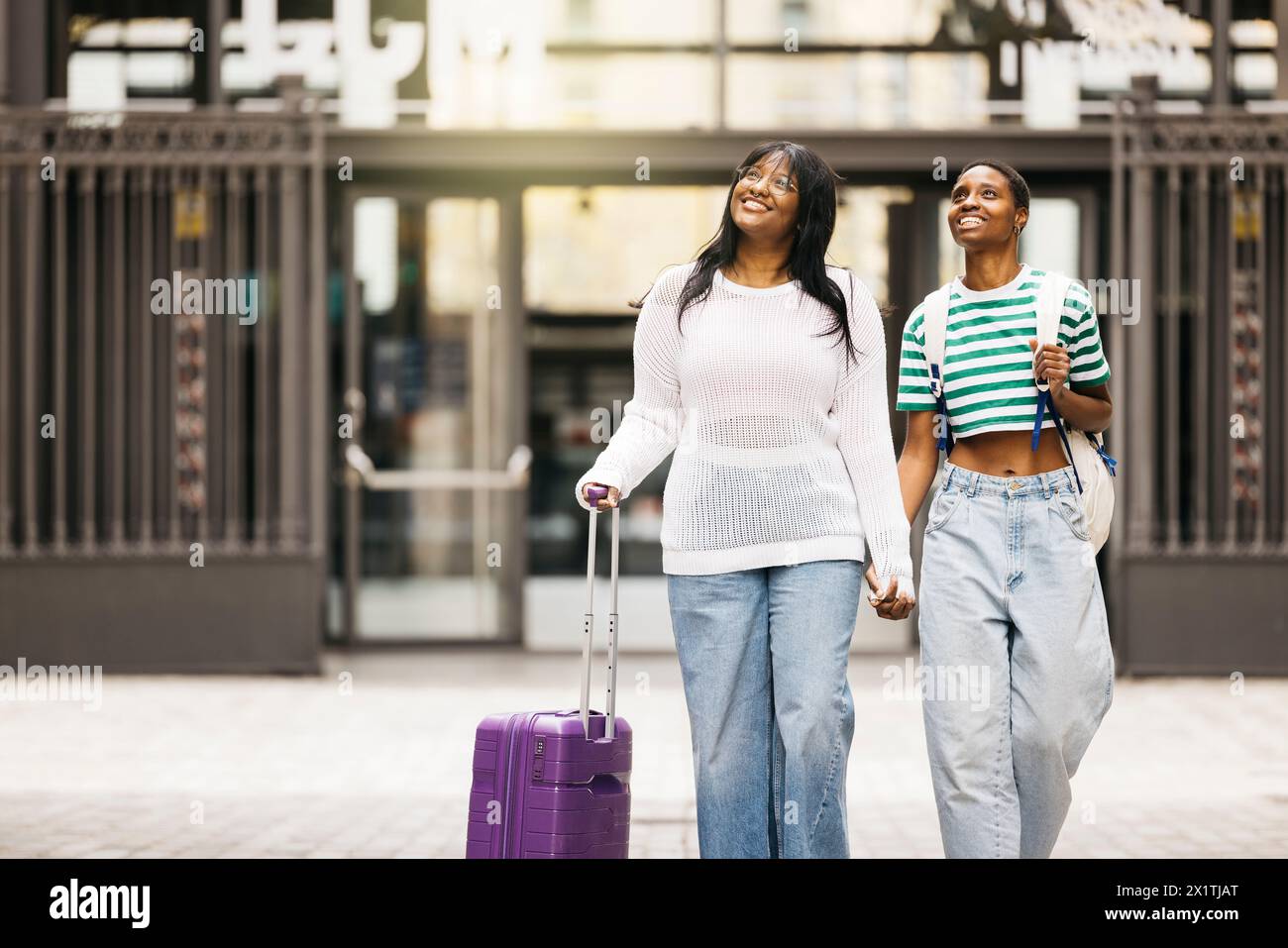 Deux jeunes femmes joyeuses tirant une valise et marchant ensemble à travers une ville pendant un week-end. Banque D'Images