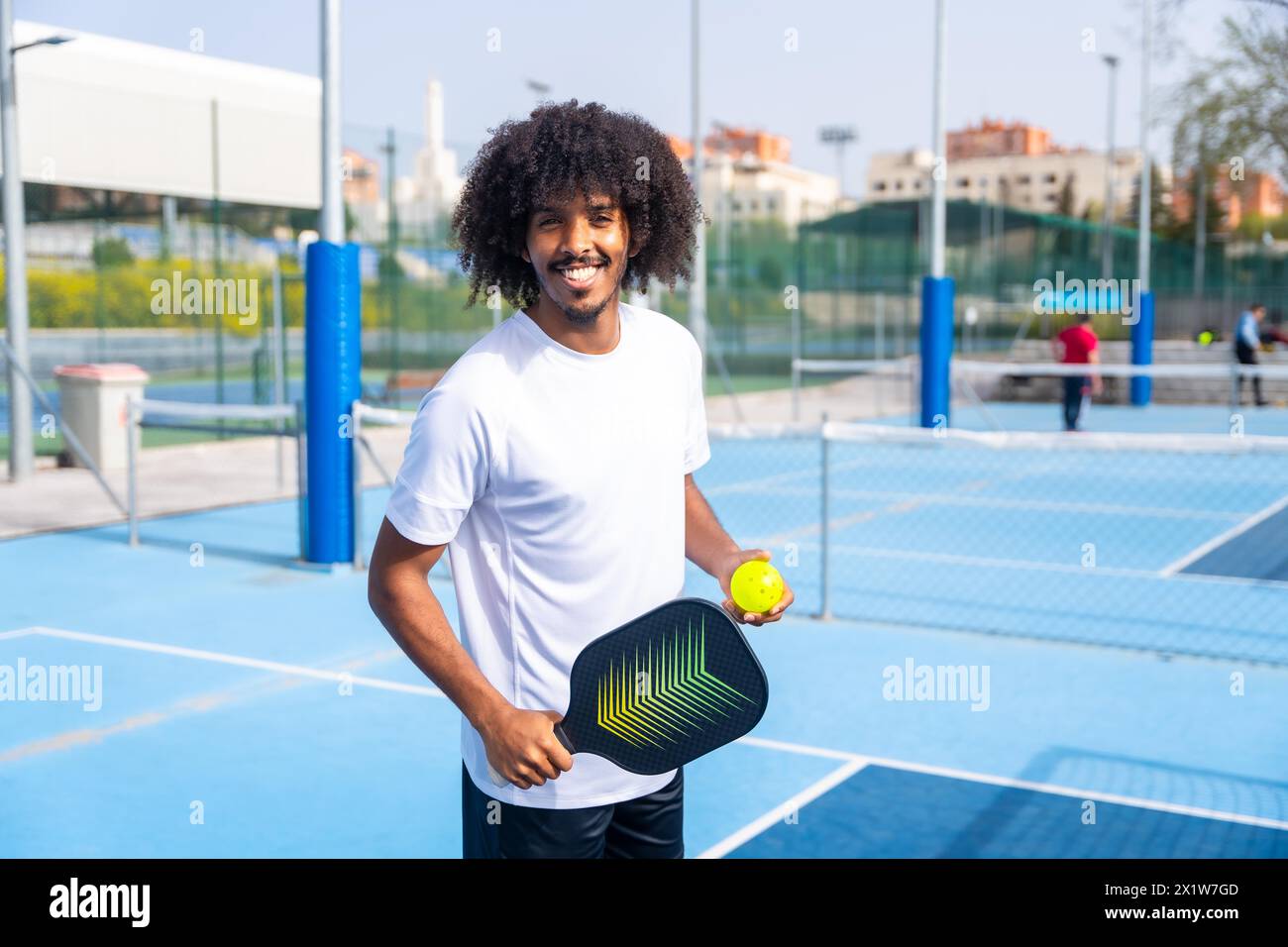 Portrait avec un homme à la coiffure afro souriant dans un terrain de pickleball tenant une raquette et une balle Banque D'Images