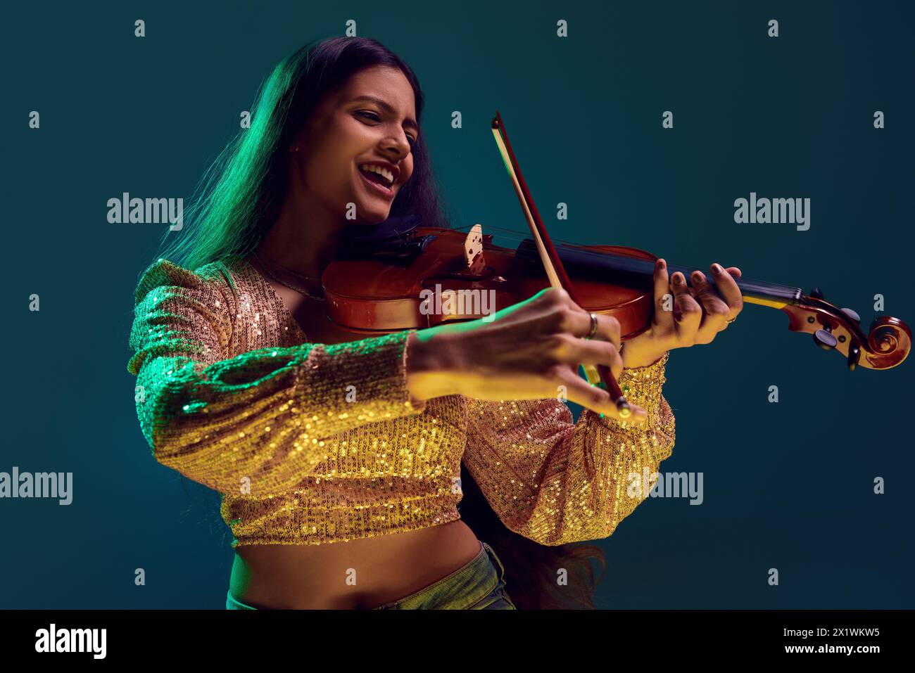 Heureuse jeune femme indienne, violoniste en top or étincelant, immergée dans la musique au néon sur fond dégradé. Banque D'Images