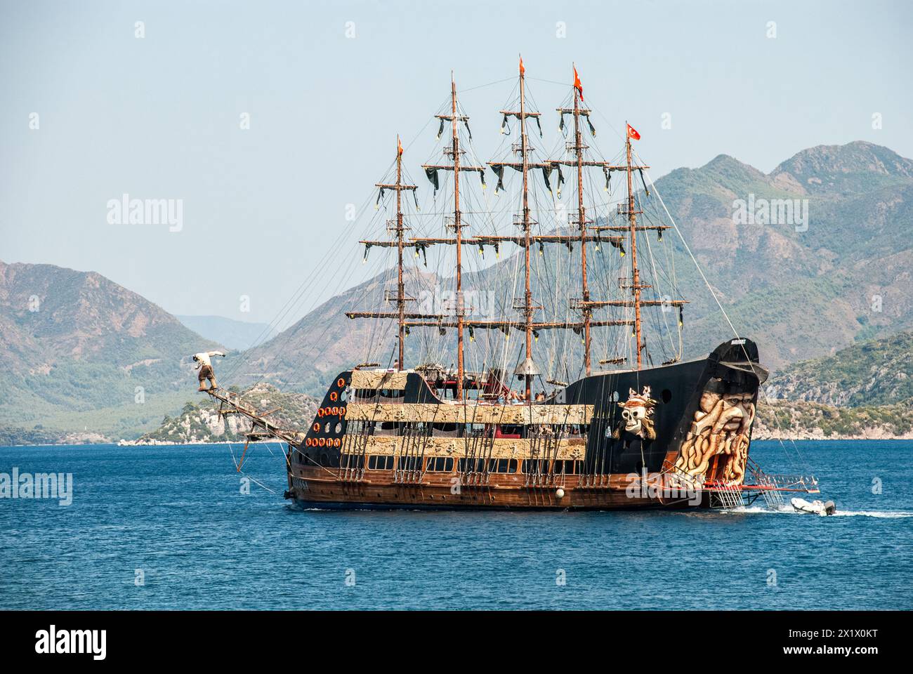 Un bateau pirate divertissant avec ses voiles noires abaissées, transportant des touristes s'amusant à bord, navigue dans un port tranquille entouré de collines verdoyantes Banque D'Images