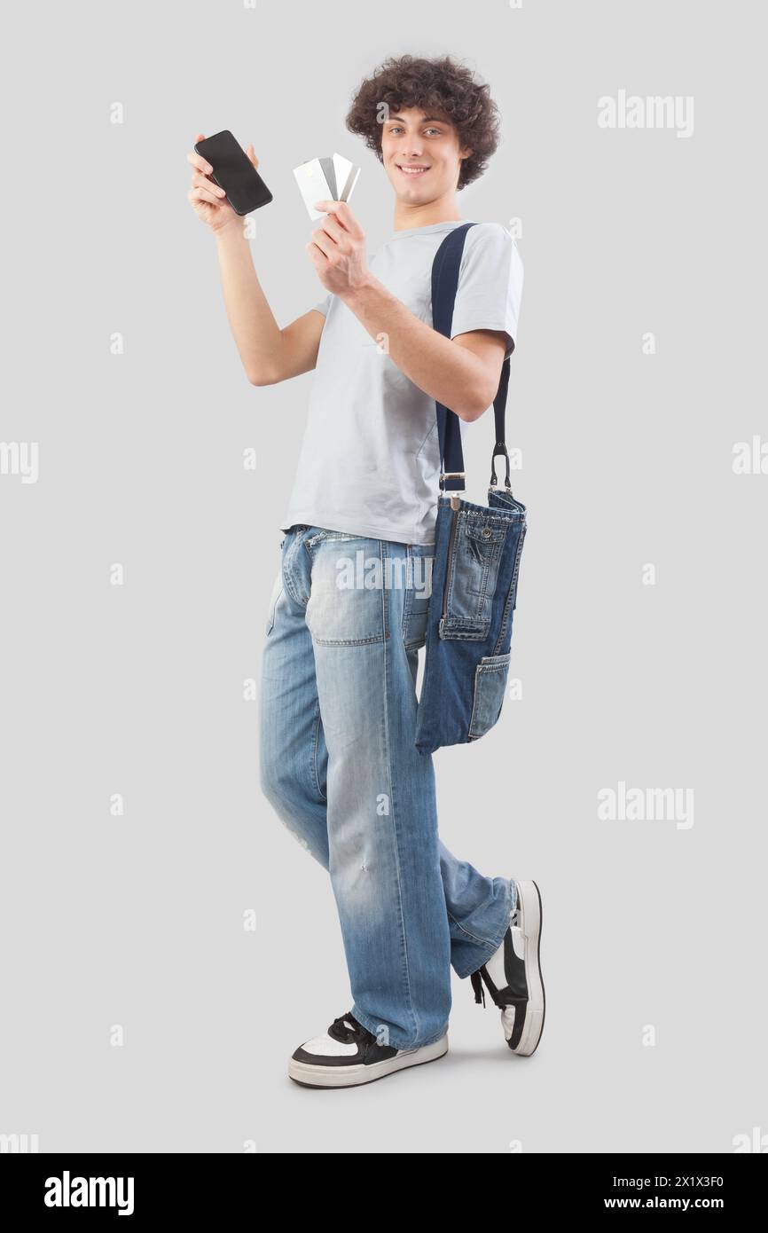 jeune homme souriant et beau montrant téléphone portable et carte de crédit habillé en jean et t-shirt avec sac à bandoulière, isolé sur fond gris Banque D'Images