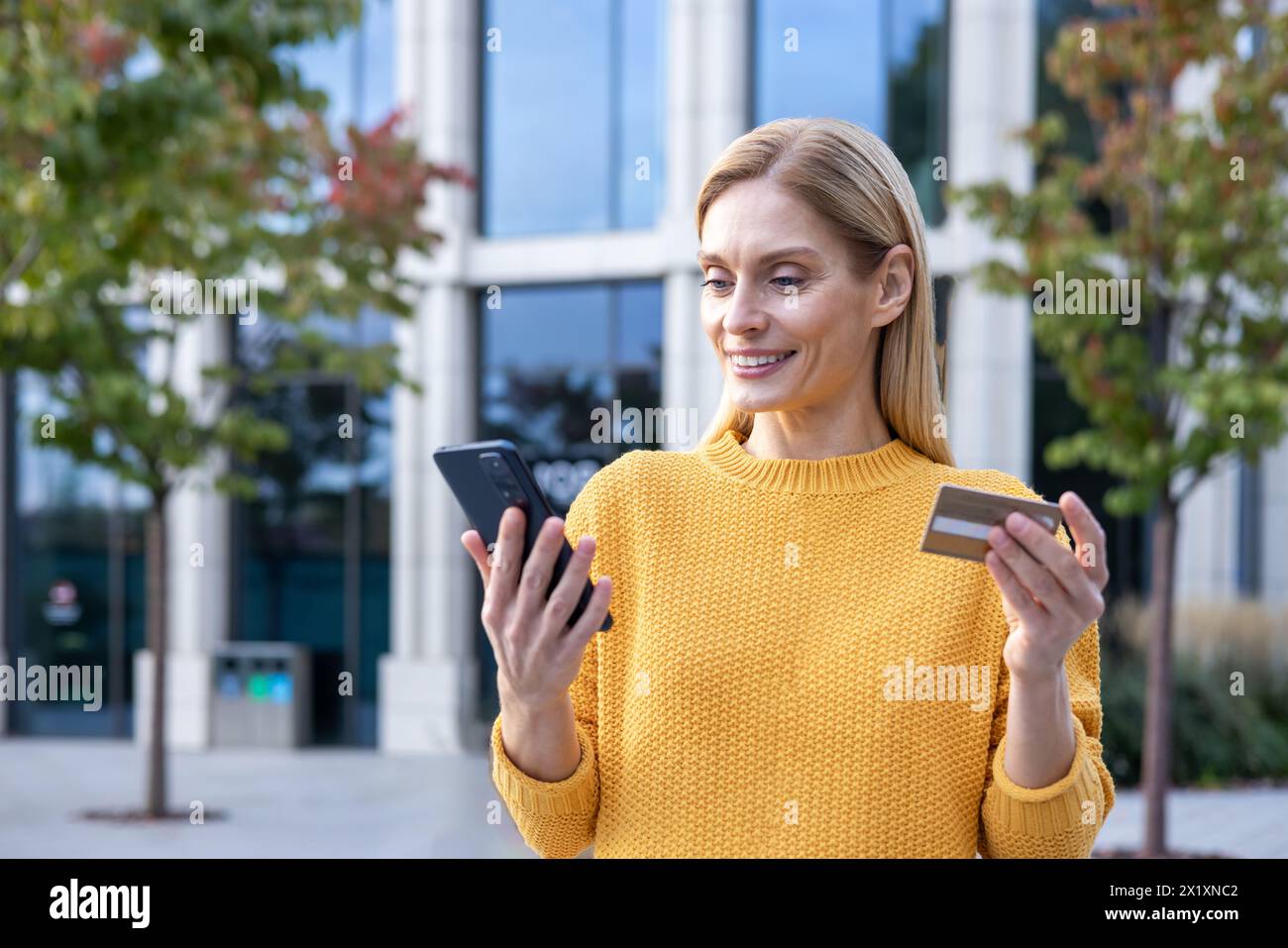 Femme blonde joyeuse en pull jaune à l'aide d'un smartphone et tenant une carte de crédit à l'extérieur, peut-être magasiner ou gérer les finances en ligne. Banque D'Images