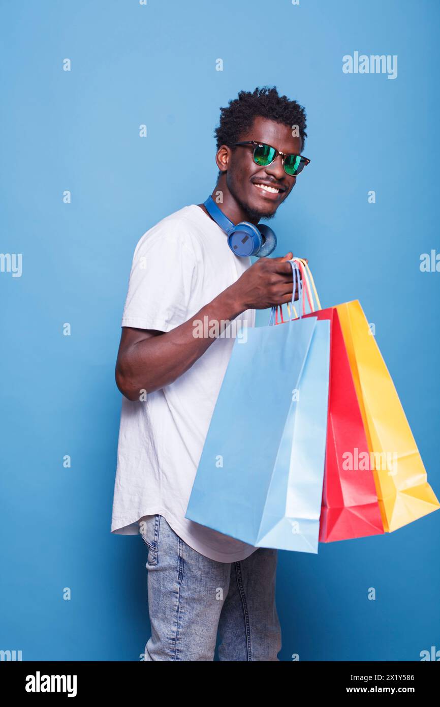 Gars afro-américain portant des lunettes de soleil, tenant des sacs à provisions, et debout devant un fond bleu isolé. Homme noir avec casque sans fil sur le cou, transportant des colis colorés. Banque D'Images