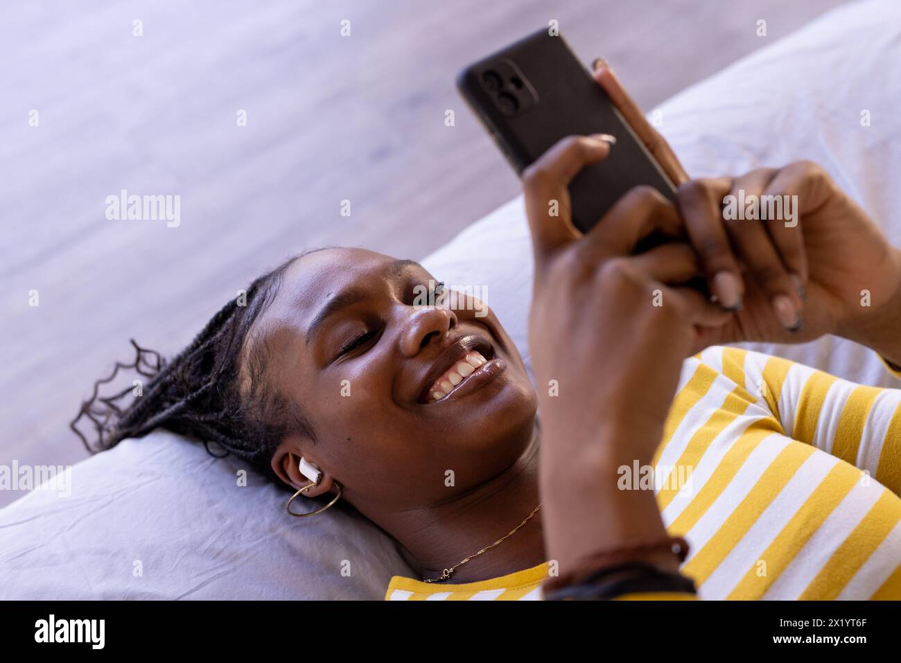 Jeune femme afro-américaine couchée sur le lit à la maison, tenant un smartphone, espace de copie, à la maison. Elle a les cheveux tressés foncés, portant une rayure jaune Banque D'Images