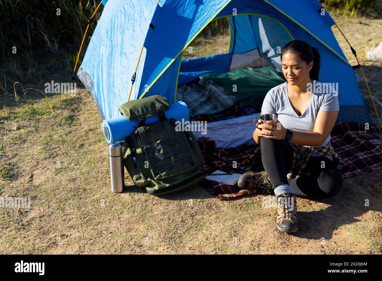 Randonneur féminin biracial tenant une tasse est assis près d'une tente. Elle a les cheveux bruns, les yeux foncés, et porte des vêtements de plein air décontractés, inaltérés, nature d'aventure. Banque D'Images