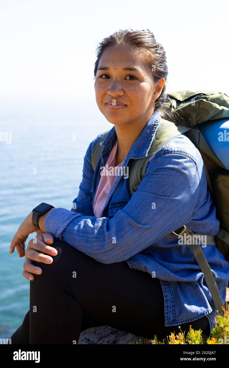 Randonneur féminin biracial assis, portant un sac à dos, aventure dans la nature Banque D'Images