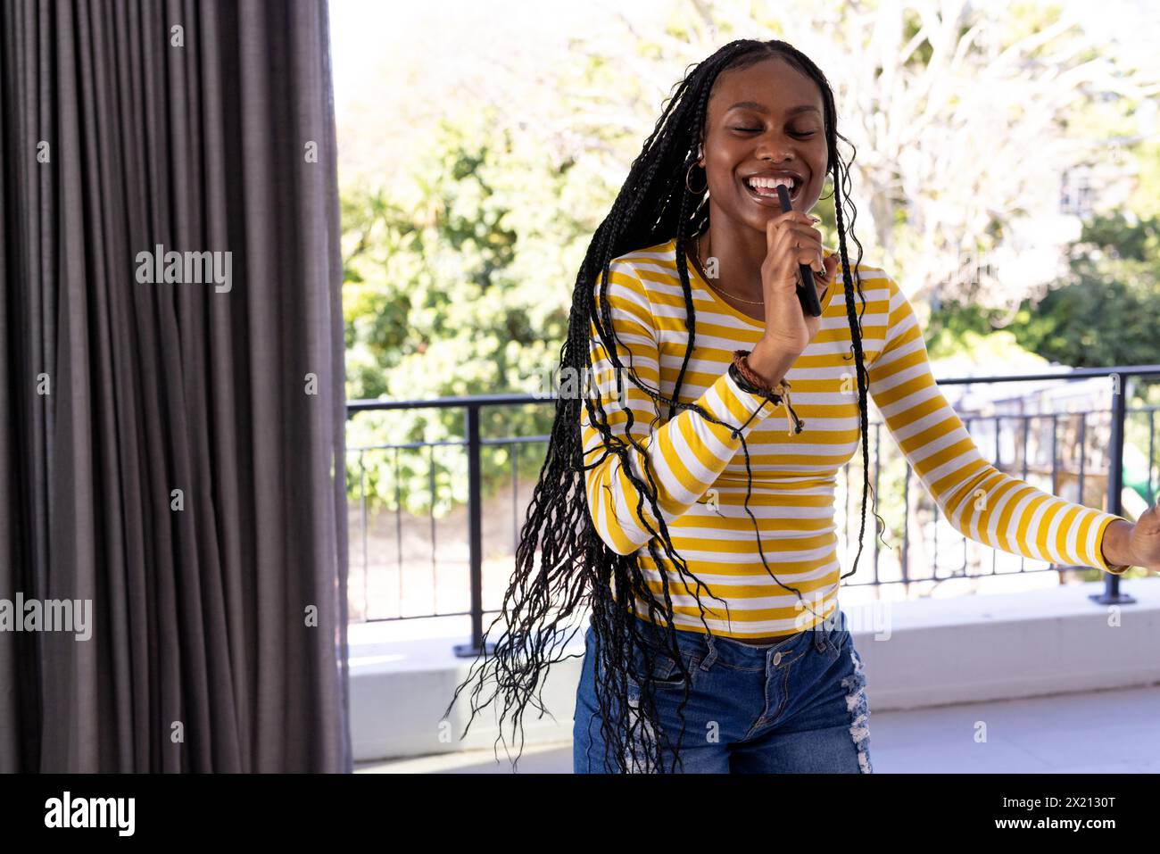 Une jeune femme afro-américaine rit à l'intérieur, debout près d'une fenêtre, un espace de copie, à la maison. Elle a de longs cheveux tressés, portant une chemise rayée, en Banque D'Images