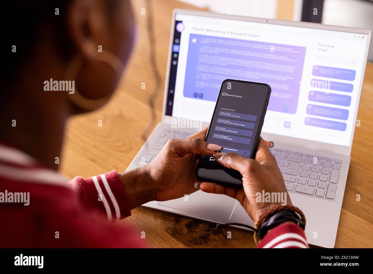 Jeune femme afro-américaine à l'intérieur avec téléphone, ordinateur portable montrant ai Chat Bot Screen à la maison Banque D'Images