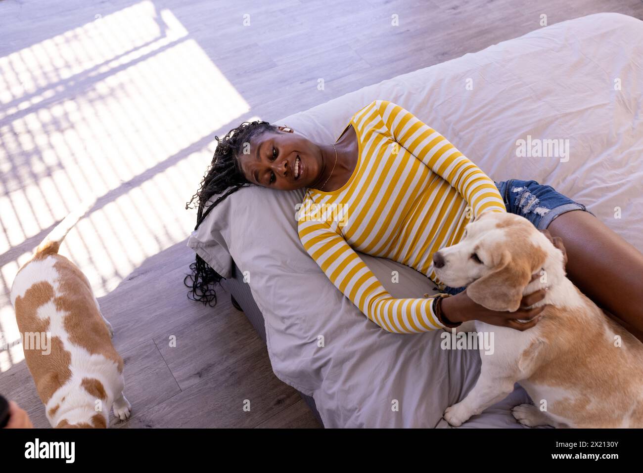 Jeune femme afro-américaine allongée sur le sol à l'intérieur à la maison, caressant un chien, copiez l'espace. Elle a les cheveux foncés, portant un haut rayé jaune et un denim Banque D'Images