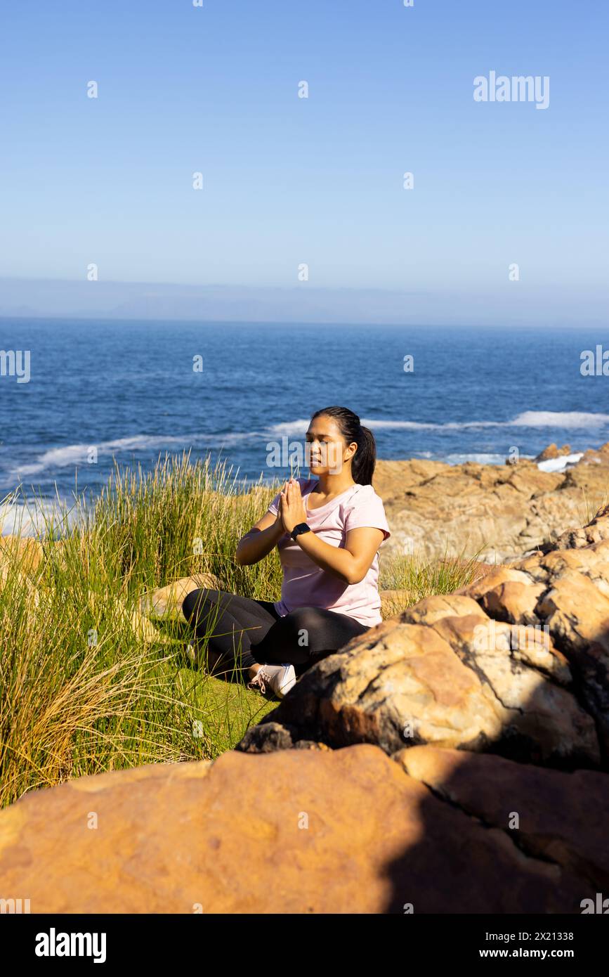 Une randonneuse biraciale profitant du yoga au bord de la mer, assise sur des rochers, espace de copie. Elle a les cheveux foncés, porte des vêtements de sport décontractés, et semble paisible, u Banque D'Images