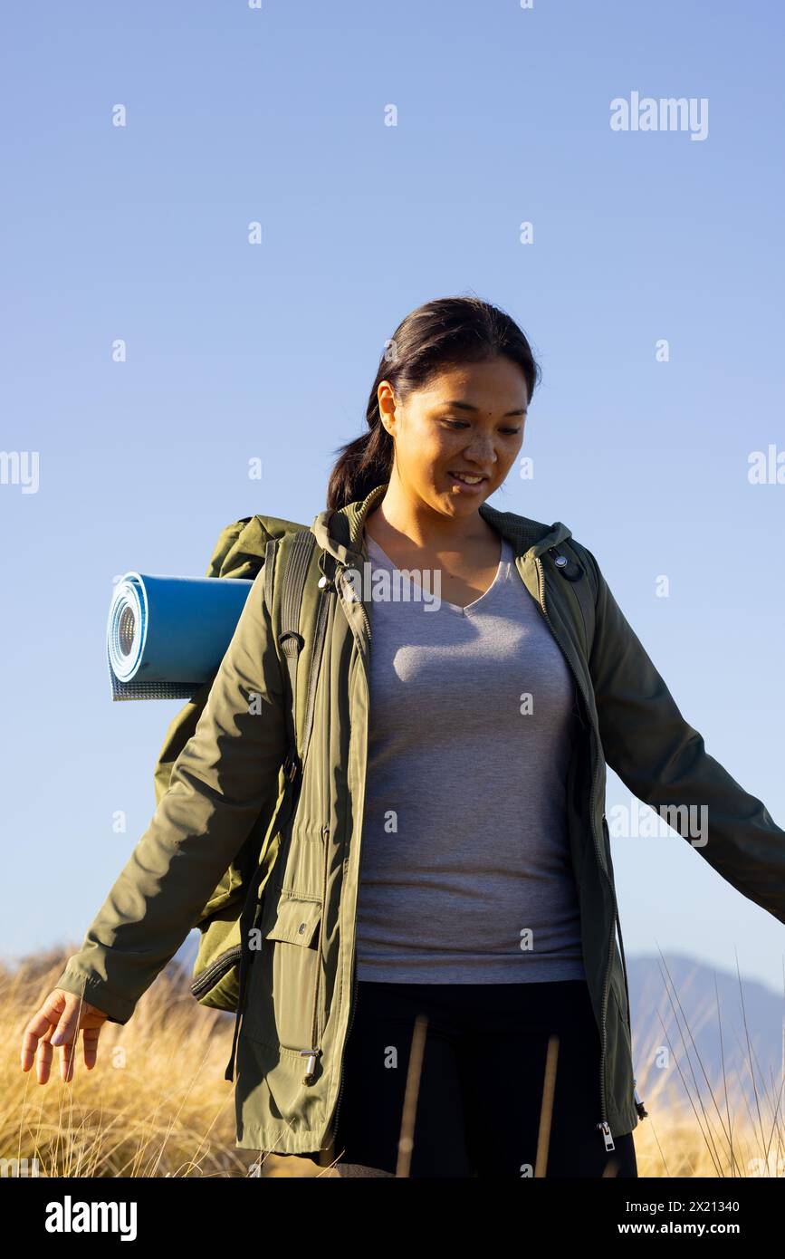 Randonneur féminin biracial avec les cheveux foncés profitant de la randonnée en montagne, espace de copie. Elle porte une veste verte, portant un tapis bleu, et a l'air détendue, inaltérable Banque D'Images