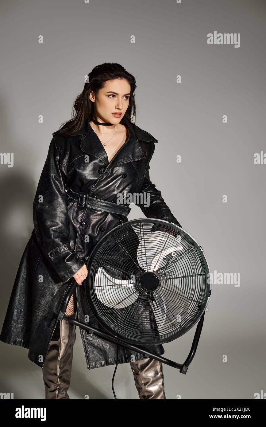 Une belle femme grande taille posant dans un manteau en cuir à la mode, tenant un ventilateur, exsudant la confiance et le style sur une toile de fond grise. Banque D'Images