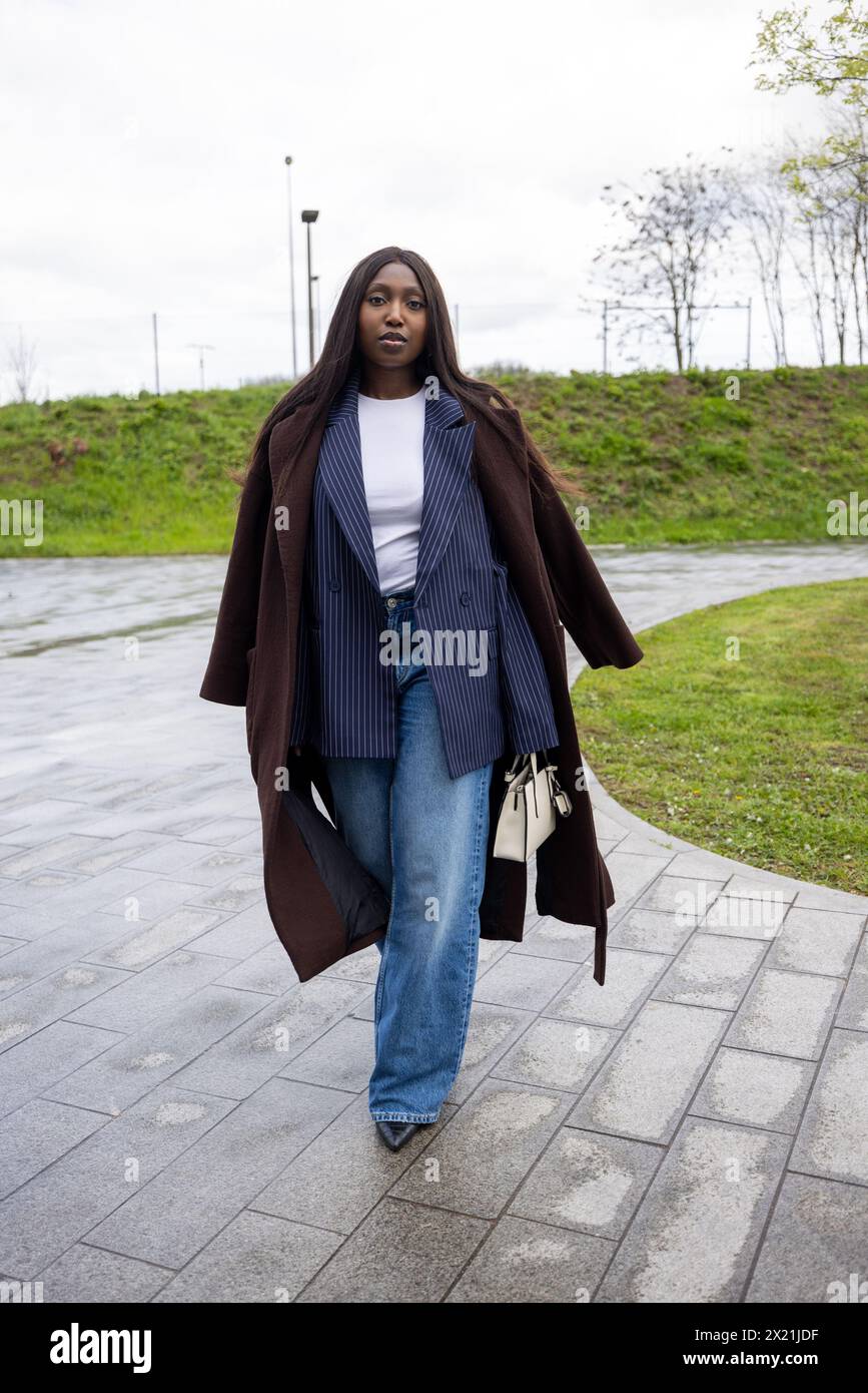 Cette photo captivante représente une jeune femme noire se promenant vigoureusement dans un parc urbain. Elle a un ensemble d'automne à plusieurs couches, avec un superbe blazer à fines rayures sous un manteau brun ouvert et riche. Son haut blanc, son Jean bleu classique et son sac à main élégant complètent le look, incarnant un mélange de praticité et de sensibilité à la mode. Dynamic Urban Walk : femme avant-gardiste en vêtements d'automne superposés. Photo de haute qualité Banque D'Images