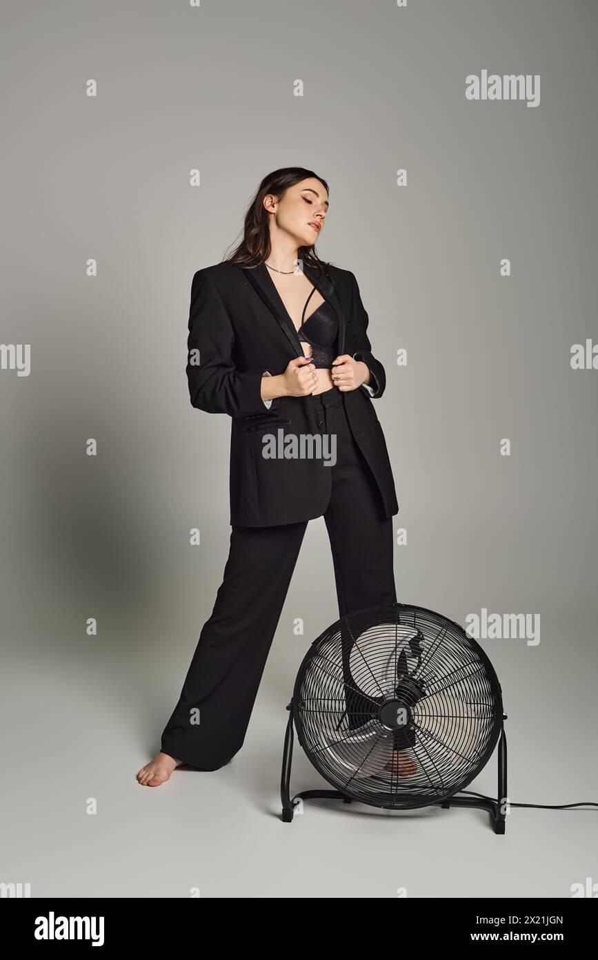 Une femme de grande taille élégante respire la confiance dans un costume sur mesure, debout gracieusement à côté d'un ventilateur en rotation sur un fond gris. Banque D'Images