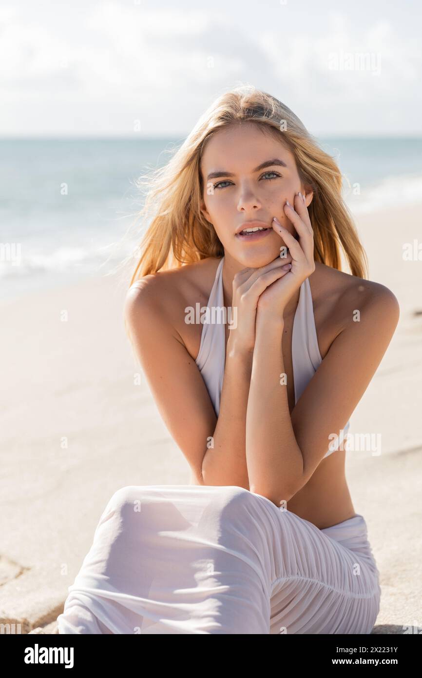 Une jeune femme blonde prend la pose assise sur les rives sablonneuses de Miami Beach, dégageant sérénité et beauté. Banque D'Images