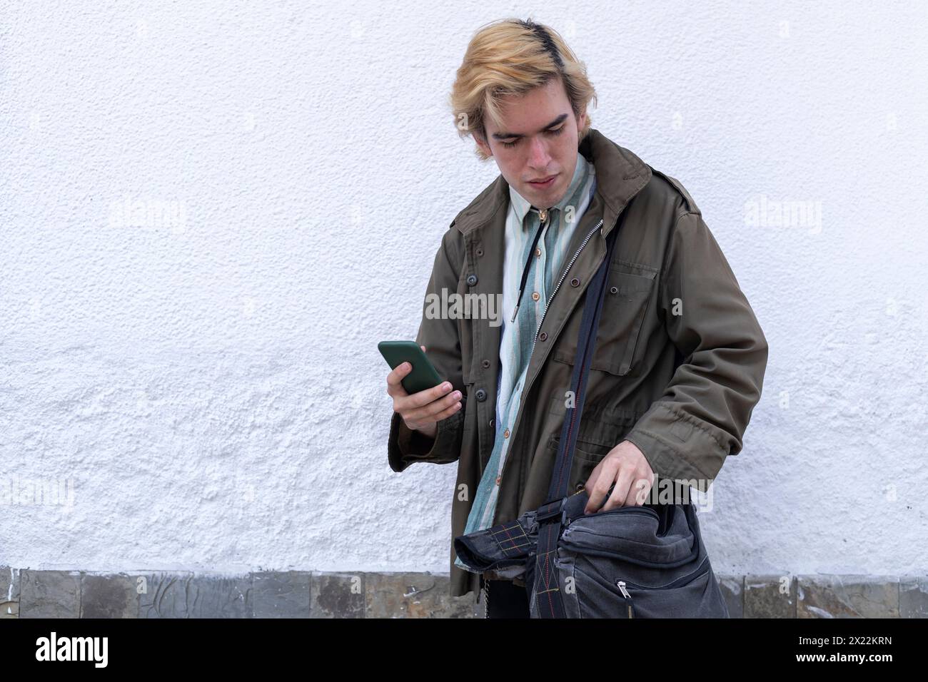 Jeune homme latino-américain dans la rue vérifiant son sac à bandoulière, tenant un téléphone portable Banque D'Images