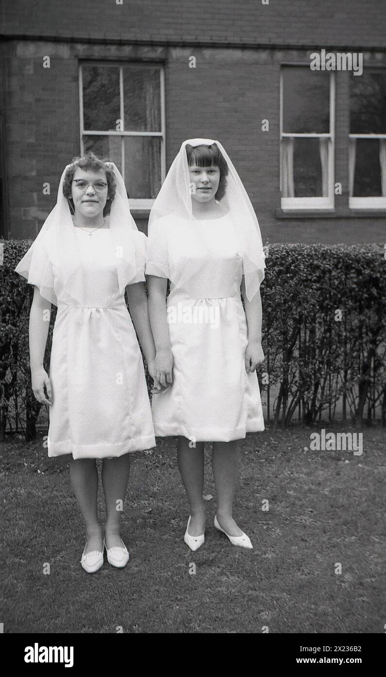 Années 1970, historique, deux écolières catholiques en robes blanches et portant des voiles de cheveux, catholiques debout pour une photo après avoir eu leur première sainte communion, Angleterre, Royaume-Uni. Banque D'Images