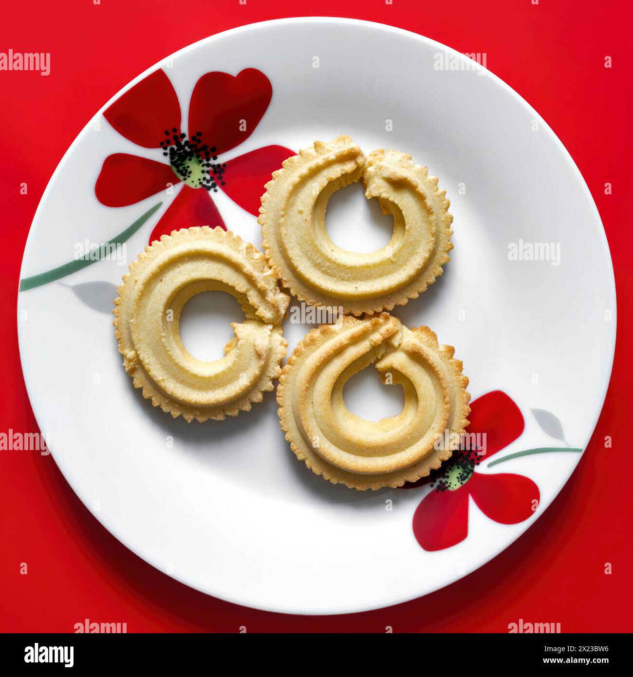 Trois biscuits vanille viennois Whirls sur plaque blanche avec motif de fleur rouge, pris sur un fond rouge Banque D'Images