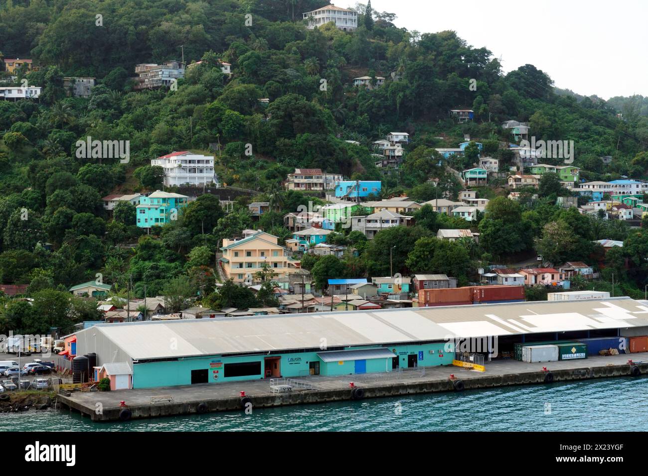 Maisons colorées typiques dans le style des Caraïbes construites sur une colline pleine de végétation verte observée depuis le port de chargement de conteneurs. Banque D'Images
