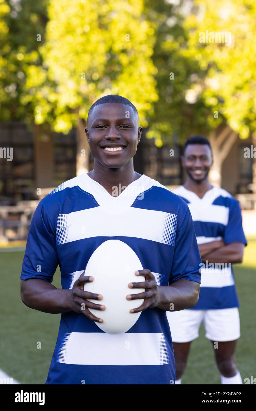 Deux jeunes athlètes afro-américains tiennent un ballon de rugby, souriant, sur un terrain en plein air Banque D'Images
