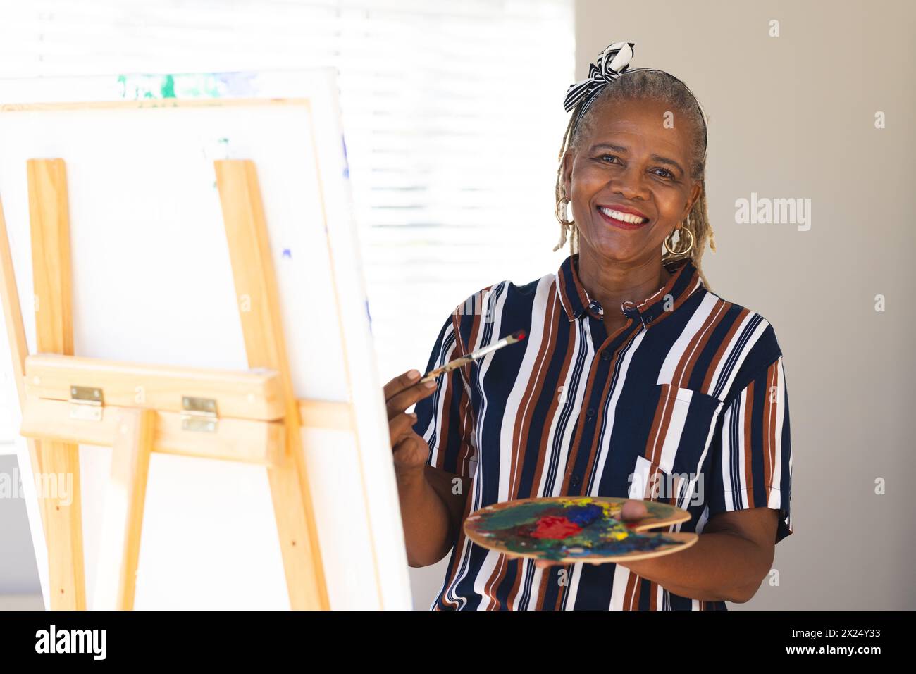 Femme aînée afro-américaine peignant sur toile à la maison, portant une chemise rayée. Elle a les cheveux gris, souriant, tenant la palette et le pinceau, inaltérés Banque D'Images