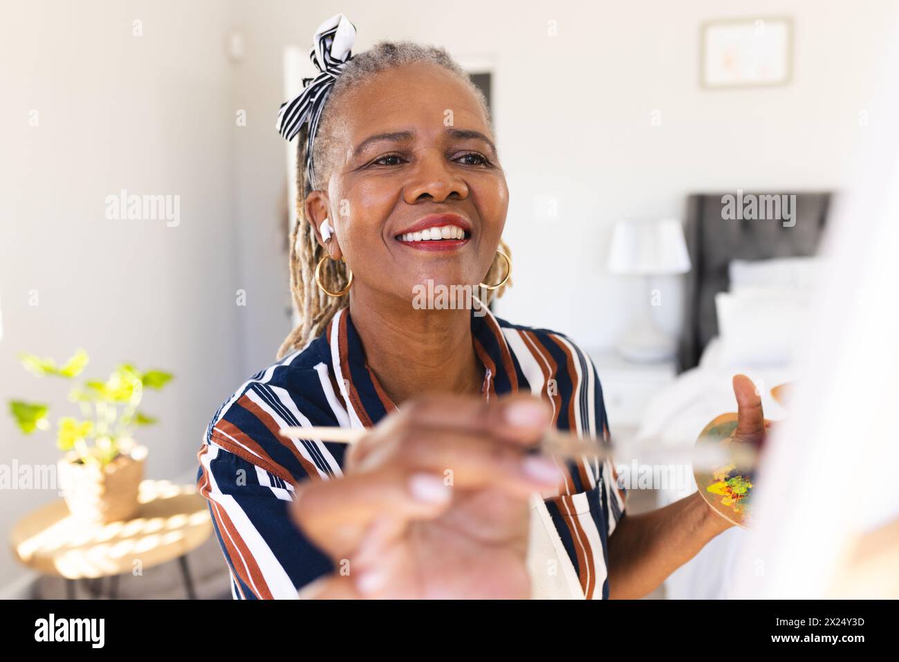 Une femme afro-américaine senior peint sur toile à la maison, souriant joyeusement. Elle a des dreadlocks gris, porte une chemise rayée, et un bandeau, unalt Banque D'Images