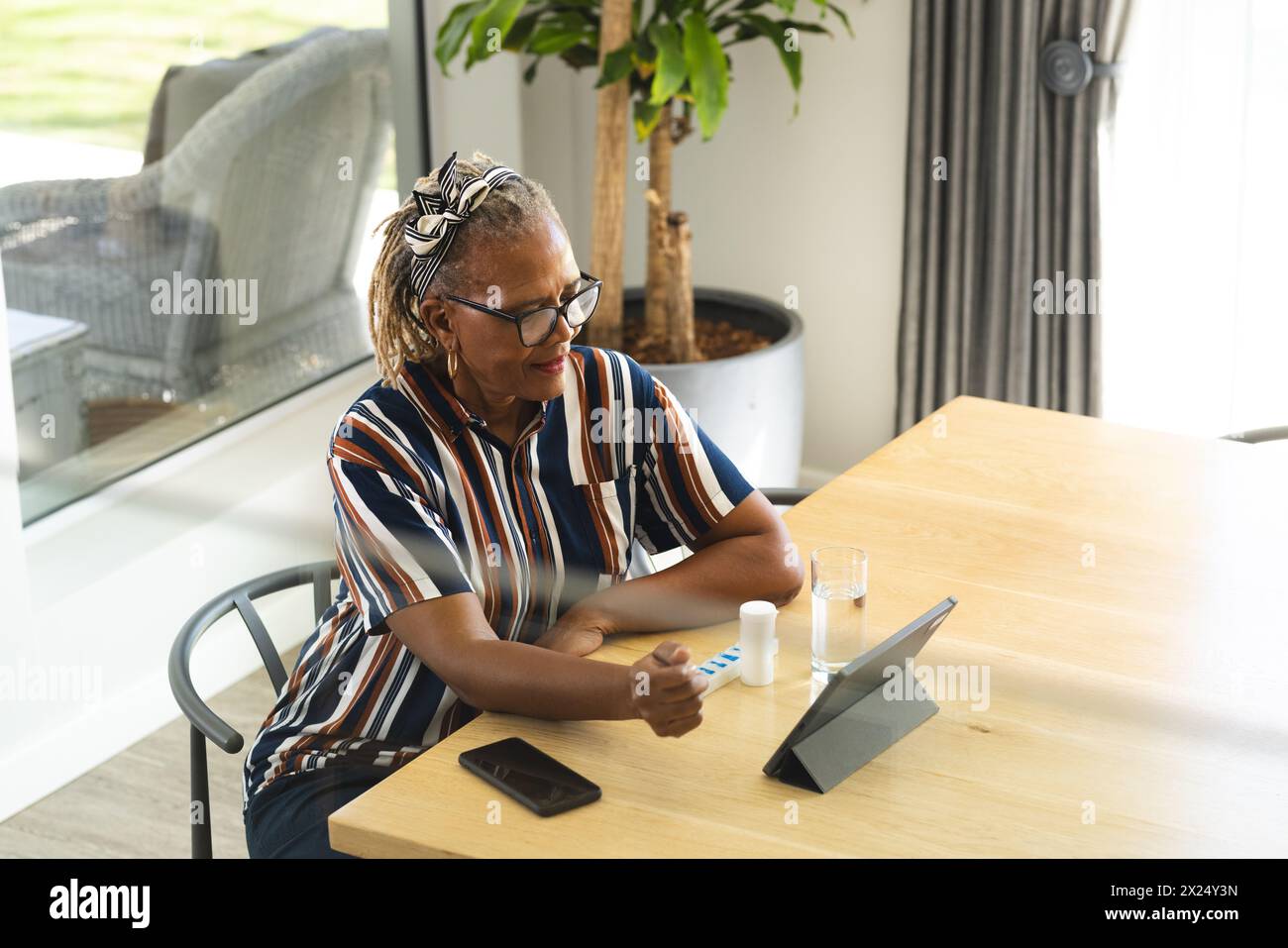 Femme aînée afro-américaine assise à la maison à table, tenant des médicaments et utilisant une tablette Banque D'Images