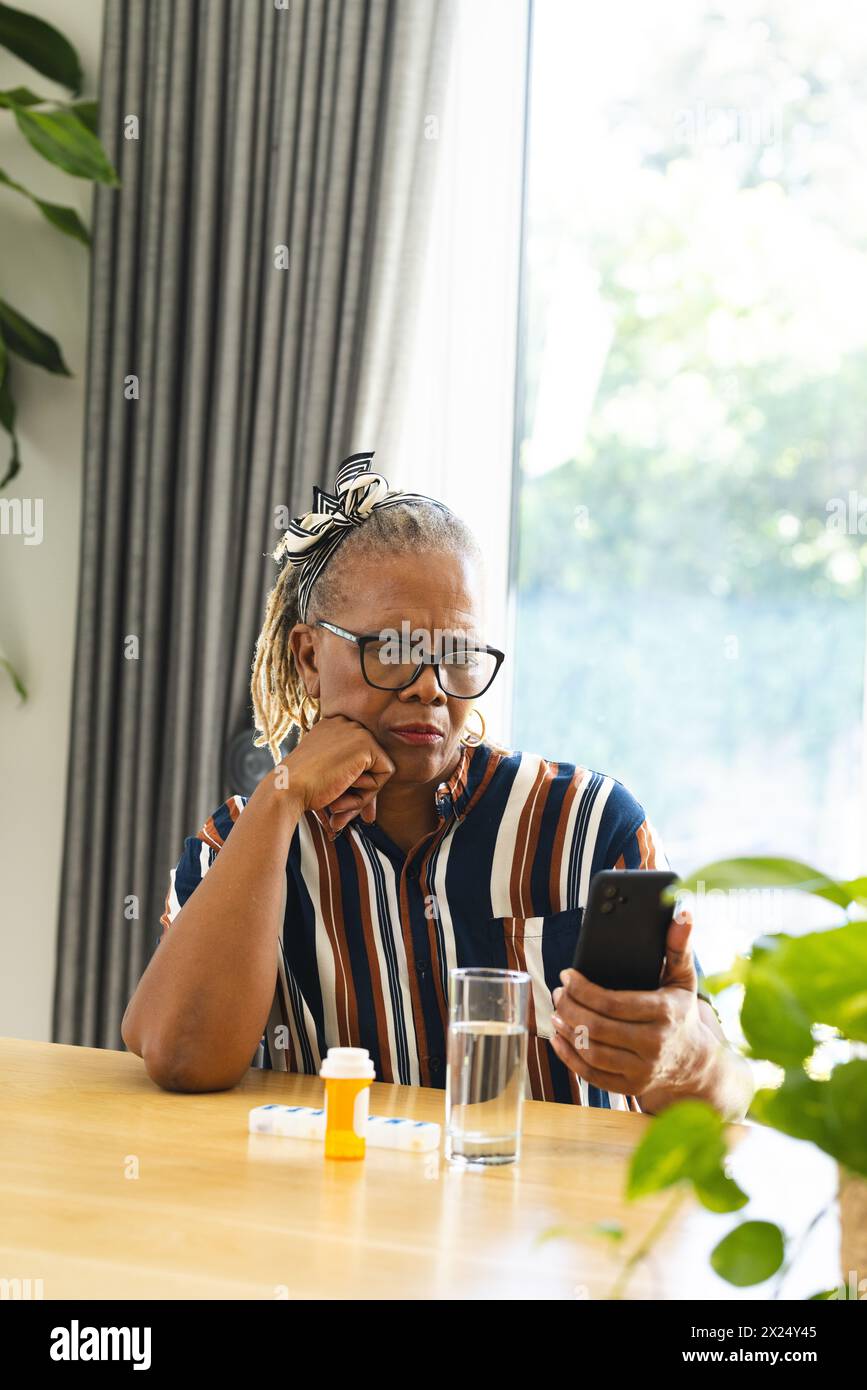 Femme aînée afro-américaine portant des lunettes regardant un smartphone à la maison et prenant des médicaments Banque D'Images