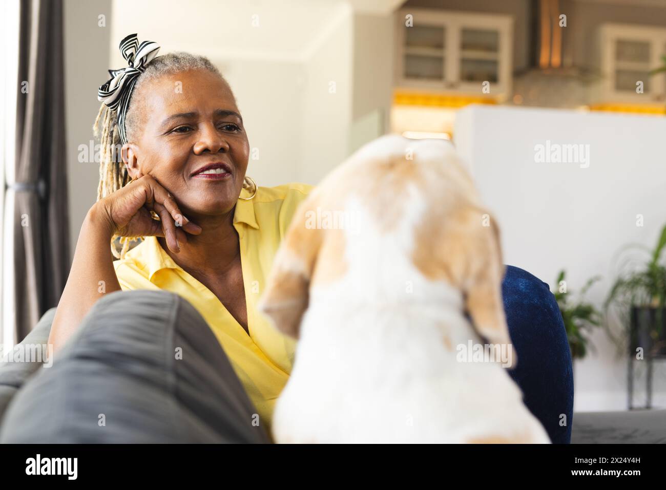 Une femme afro-américaine séniore se détache à la maison avec son chien. Elle a les cheveux gris, porte une chemise jaune, et sourit doucement. Banque D'Images