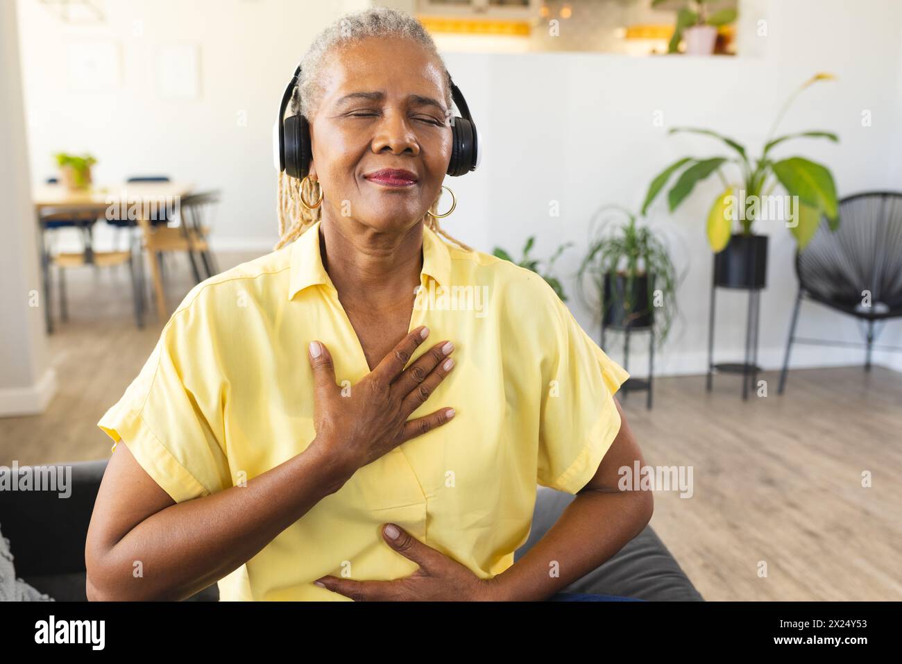 Une femme afro-américaine senior appréciant la musique avec des écouteurs à la maison, la main sur le cœur, l'espace de copie Banque D'Images